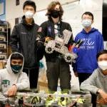 A group of five student pose for a photo with a robotics project in a classroom.